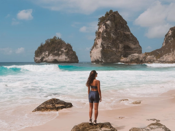 Mulher de costas para a câmera observando mar cristalino com formações rochosas grandes e levemente pontiagudas