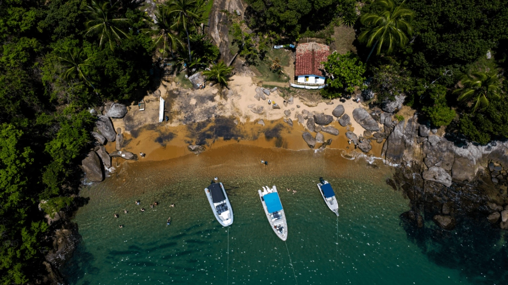 Vista aérea de praia com pequena faixa de areia, pedras e vegetação. Há três barcos estacionados sobre o mar