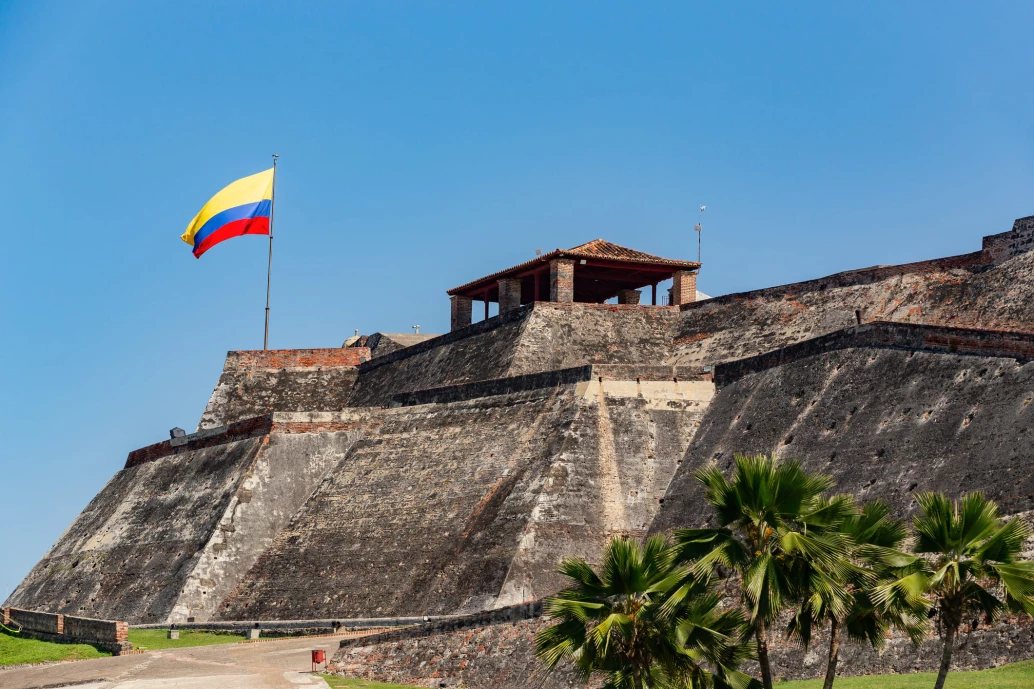 Fortaleza de um castelo na cidade de Cartagena. O muro é alto e inclinado, com um abrigo na parte superior e uma bandeira da Colômbia hasteada. À frente, algumas palmeiras se destacam na foto, que foi feita à luz do dia.