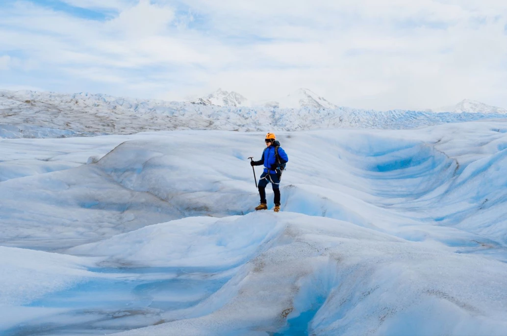 Homem com trajes de frio e equipamentos para andar sobre a geleira. Em pé, ele posa para a foto em um vasto campo de gelo, que se estende sobre um relevo ao fundo