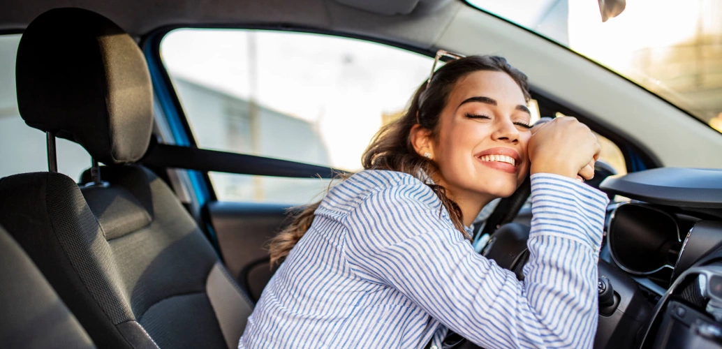 Mulher jovem sorri de olhos fechados e se debruça sobre o volante de um carro.