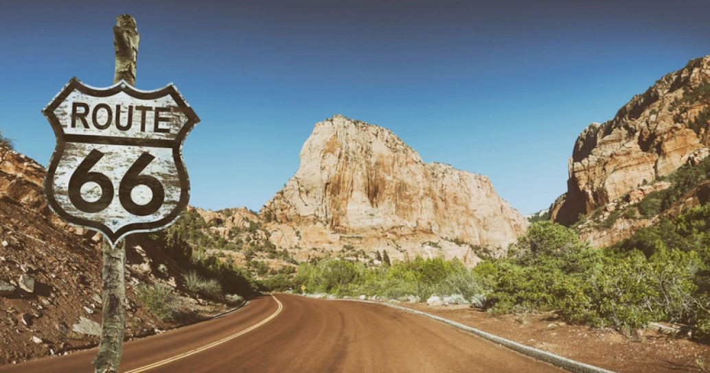 Trecho da Rota 66, nos Estados Unidos. No canto esquerdo, uma placa rústica escrito “Route 66”. Há montanhas rochosas na beira da estrada e ao fundo da imagem, além de pouca vegetação.