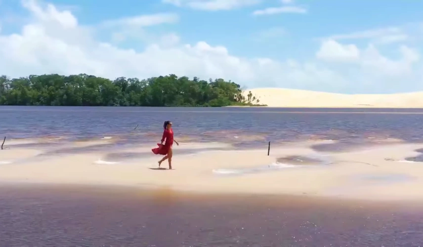Mulher de vestido vermelho caminha por lagoa de águas rasas margeada por dunas de areia branca e vegetação nativa