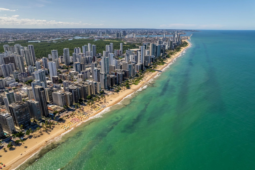 Vista aérea de praia urbana com enorme faixa de areia e grandes edifícios modernos na orla de Recife. A água do mar possui um tom esverdeado