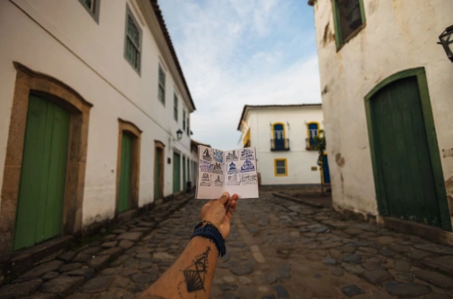 Mão de um homem segurando um passaporte com alguns carimbos. O cenário é uma rua de pedra com construções coloniais, em cidade histórica