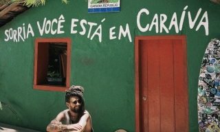Músico Gabriel Elias sentado em frente à casa de fachada na cor verde com os dizeres "sorria você está em caraíva"