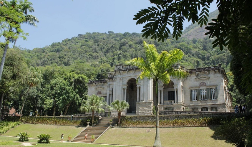 Um palácio em estilo imperial com jardim coberto de grama verde e várias árvores atrás da construção