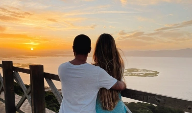Casal de costas, abraçados, assistindo ao pôr do sol em um mirante com vista para o mar.