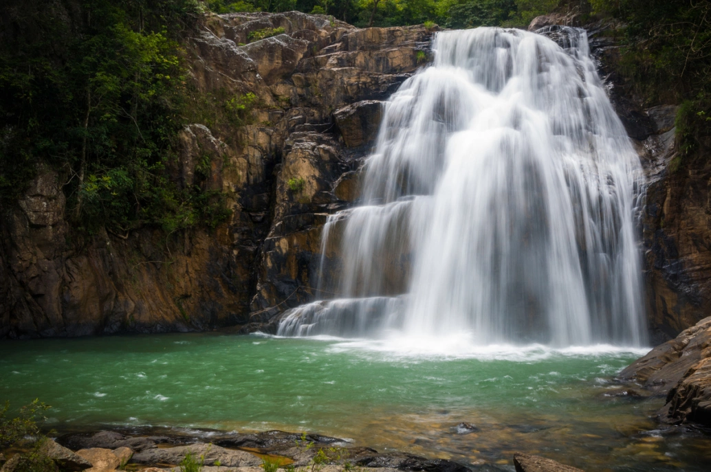 Close de cachoeira natural de tamanho médio que sai de rochas e lago que se forma abaixo, com água esverdeada.