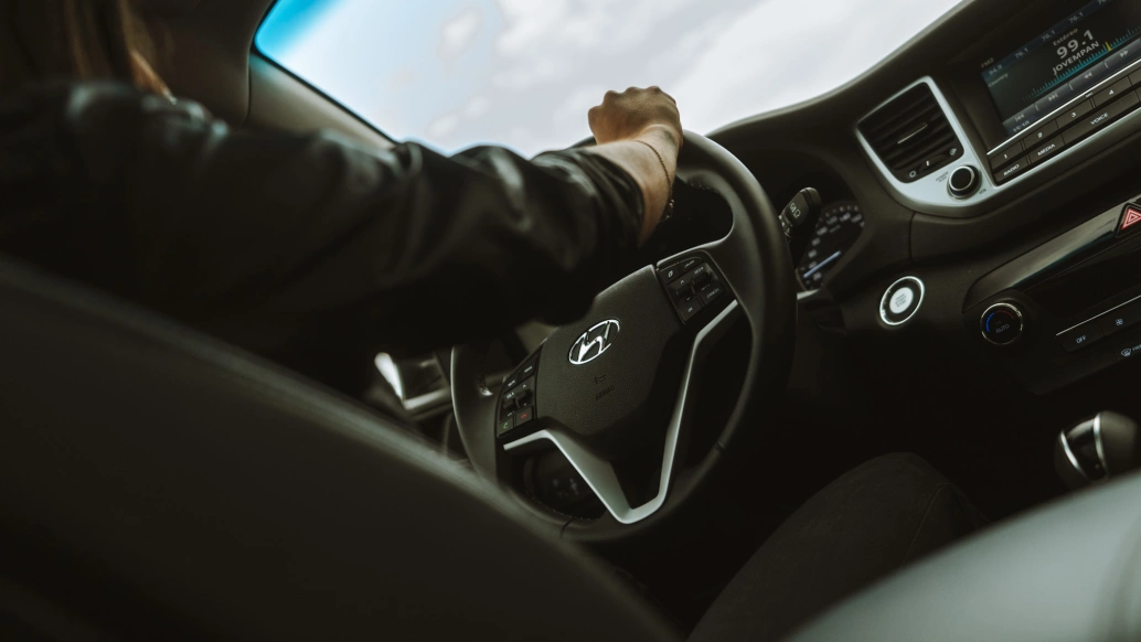 Fotografia em close do interior de um carro, focando na direção e parte do painel. Uma pessoa toca o volante.