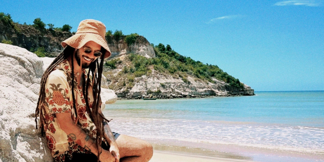 Músico Gabriel Elias se apoia em rocha em frente a praia paradisíaca em dia ensolarado