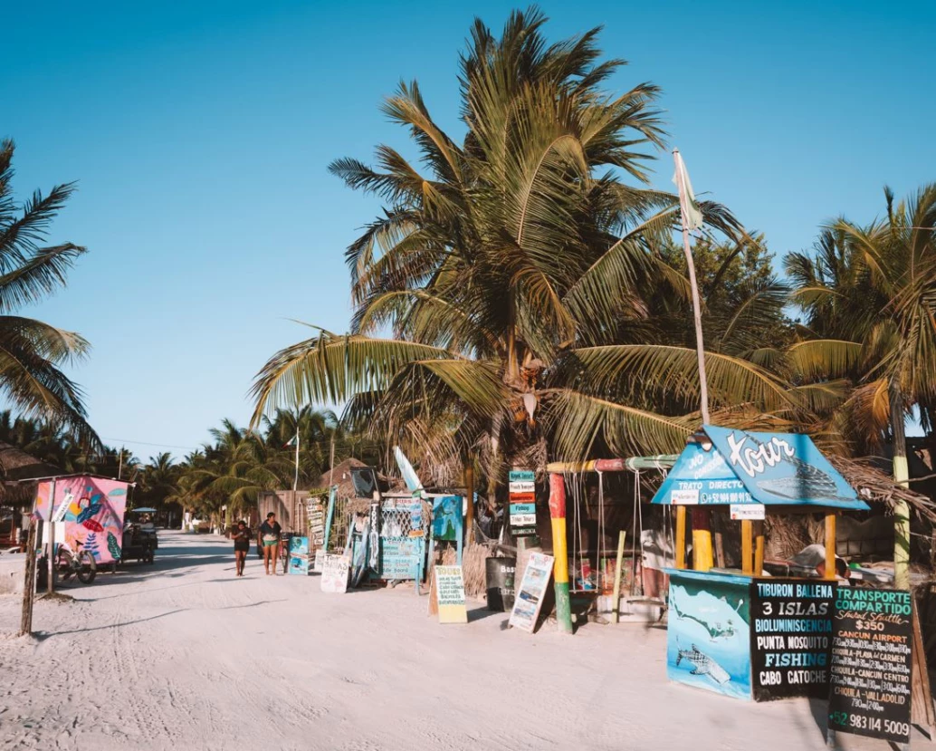 Estrada de areia avançando em paralelo a palmeiras e barracas em um dia ensolarado com céu azul, em Holbox, México.