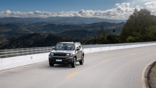 Jeep Renegade em movimento em uma estrada com montanhas verdes e um céu azul atrás.