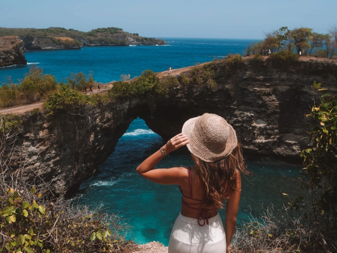 Mulher de costas para a câmera observando o oceano cortado por uma pedra com um buraco no meio, que forma uma ponte natural por cima do mar