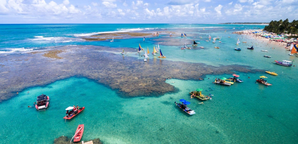 Vista aérea do mar verde-esmeralda e recifes de corais, com muitas jangadas sobre a água. Ao fundo, coqueirais, banhistas, guarda-sóis e céu azul com nuvens