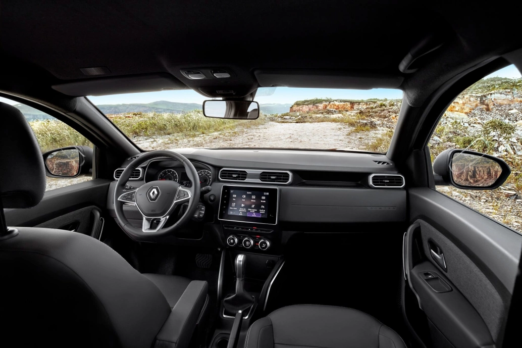 Interior da Renault Duster, mostrando volante, painel, central multimídia e parte dos bancos da frente. Através dos vidros, há uma vista para uma estrada de terra com montanhas ao fundo.