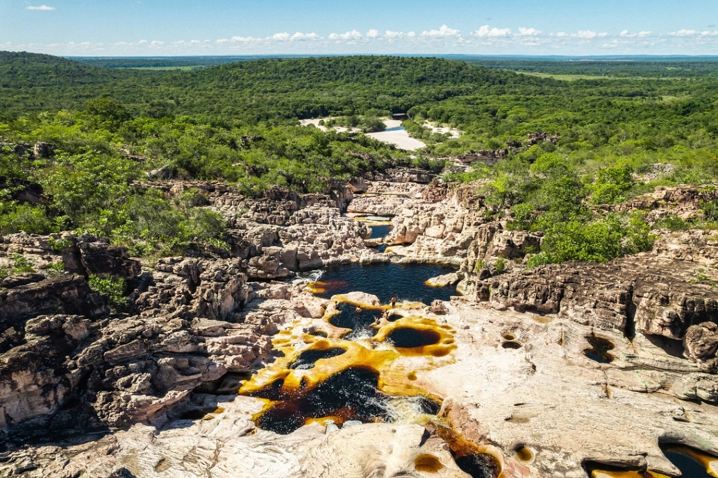 Vista panorâmica de piscinas naturais que formam poços d'água em rochas cercadas por extensa vegetação nativa com linda paisagem verde ao fundo
