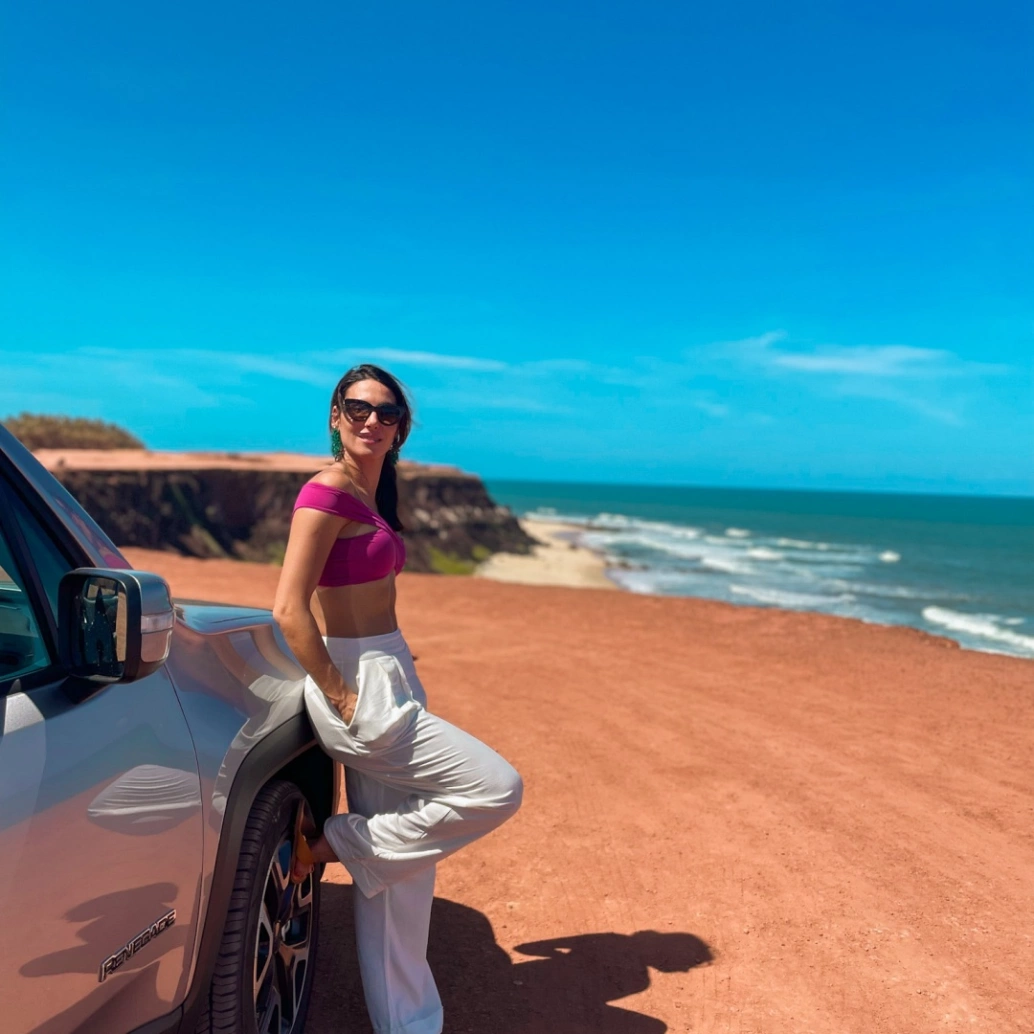 Mulher de blusa rosa e calça branca se apoia em um carro à esquerda da imagem. A vista do local são as falésias avermelhadas, o verde-esmeralda do mar e o azul do céu sem nuvens.