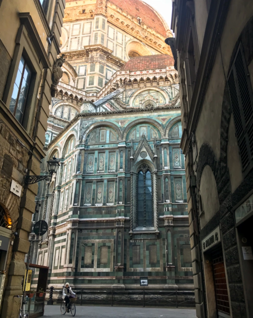 Vista imponente de uma viela da Catedral di Santa Maria del Fiori na Toscana, Itália. Destaque para sua fachada em mármore verde, branca e vermelha, com ornamentos clássicos e rebuscados, um vitral centralizado e domo, acima.