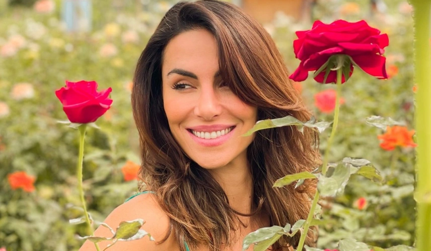 Mulher sorrindo entre duas rosas vermelhas dentro de jardim com várias plantas verdes ao fundo.