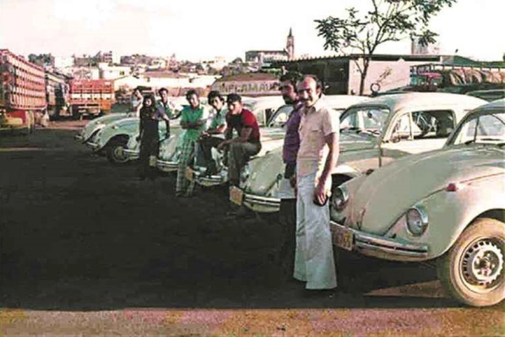 Foto da década de 1970 com seis fuscas estacionados, quatro homens em pé e três homens sentados no capô dos carros em um estacionamento.