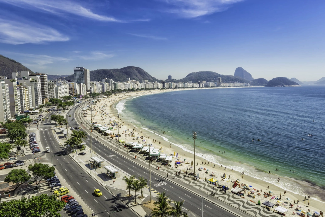 Vista aérea da Praia de Copacabana, no Rio de Janeiro. A praia termina em um calçadão. Há prédios do outro lado da avenida, e montanhas ao fundo