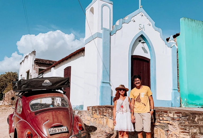Um homem e uma mulher em pé em frente a uma igreja com fachada branca em dia ensolarado com um Fusca vermelho estacionado ao lado