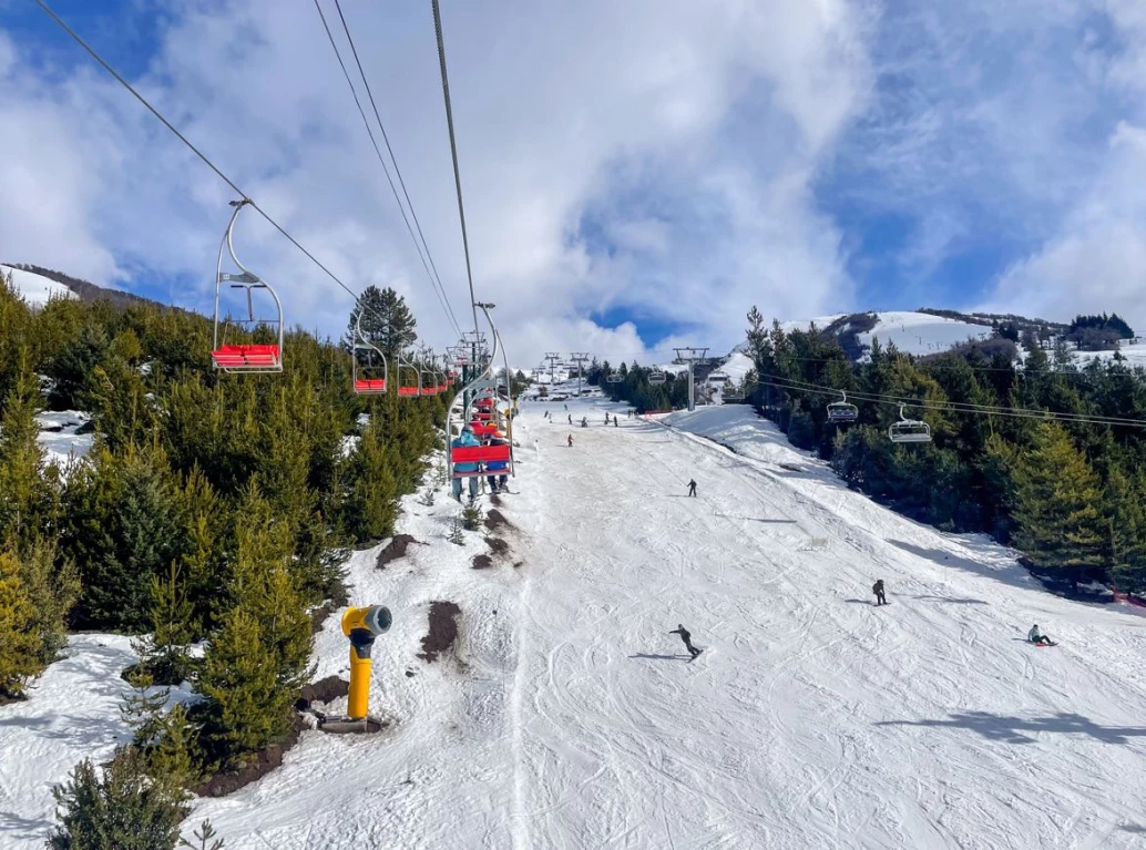 Teleféricos subindo o Cerro Catedral, em uma pista de esqui coberta por neve e margeada por árvores verdes em Bariloche, Argentina.