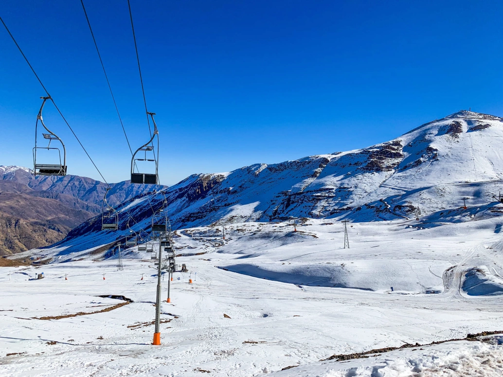 Estação de esqui em uma montanha coberta por neve. No canto esquerdo da imagem, teleférico com cadeiras individuais para transportar os praticantes da atividade
