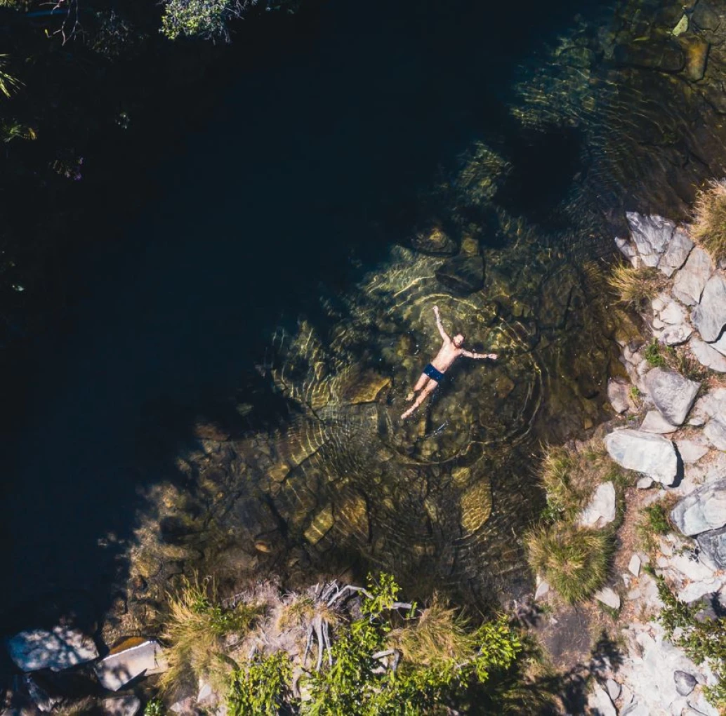 Vista aérea e distante de um homem flutuando com os braços abertos sobre o poço de uma cachoeira na Serra do Cipó. Há pedras na margem da piscina natural, além de algumas espécies de plantas rasteiras em meio a elas.