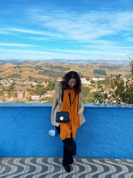 Jovem mulher trajando roupas de outono e olhando para baixo está encostada em um muro baixo de cor azul-claro e vibrante com vista panorâmica da montanhosa cidade de Águas de Lindóia, SP, ao fundo.