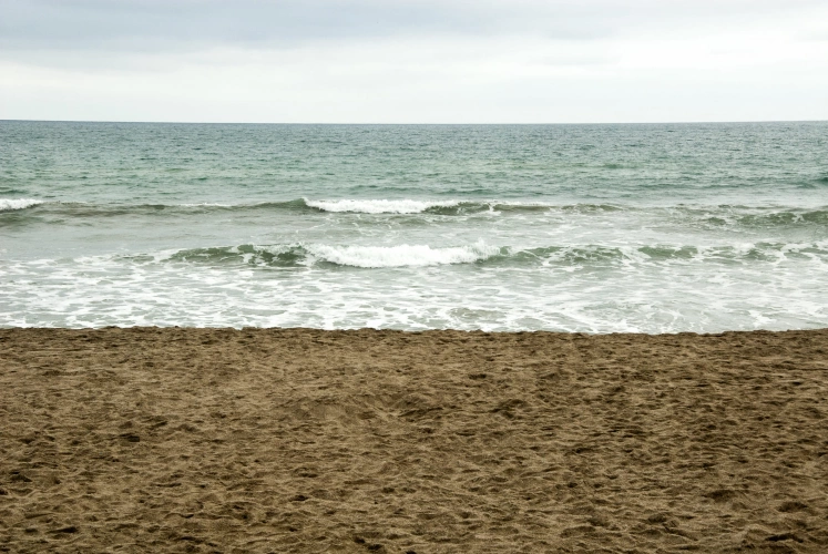 Vista frontal do mar de coloração esverdeada, com pequenas ondas. Destaque para areia de cor bege