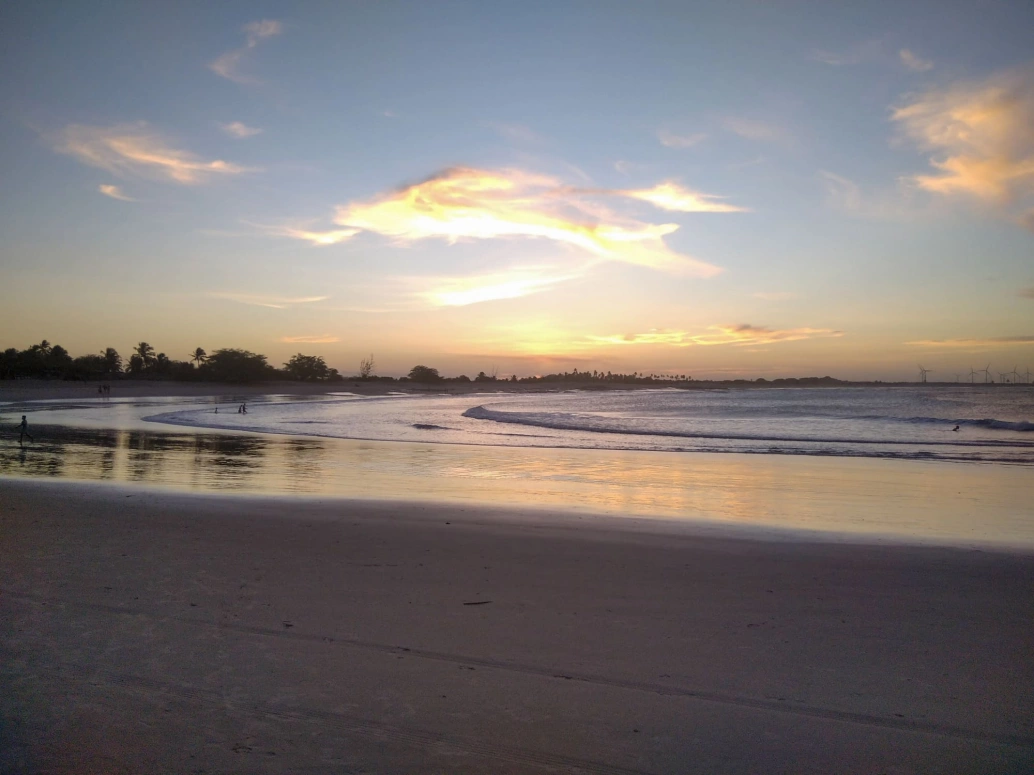 Vista de praia deserta ao cair do dia. Mares tranquilas formam um espelho do céu com poucas nuvens douradas.