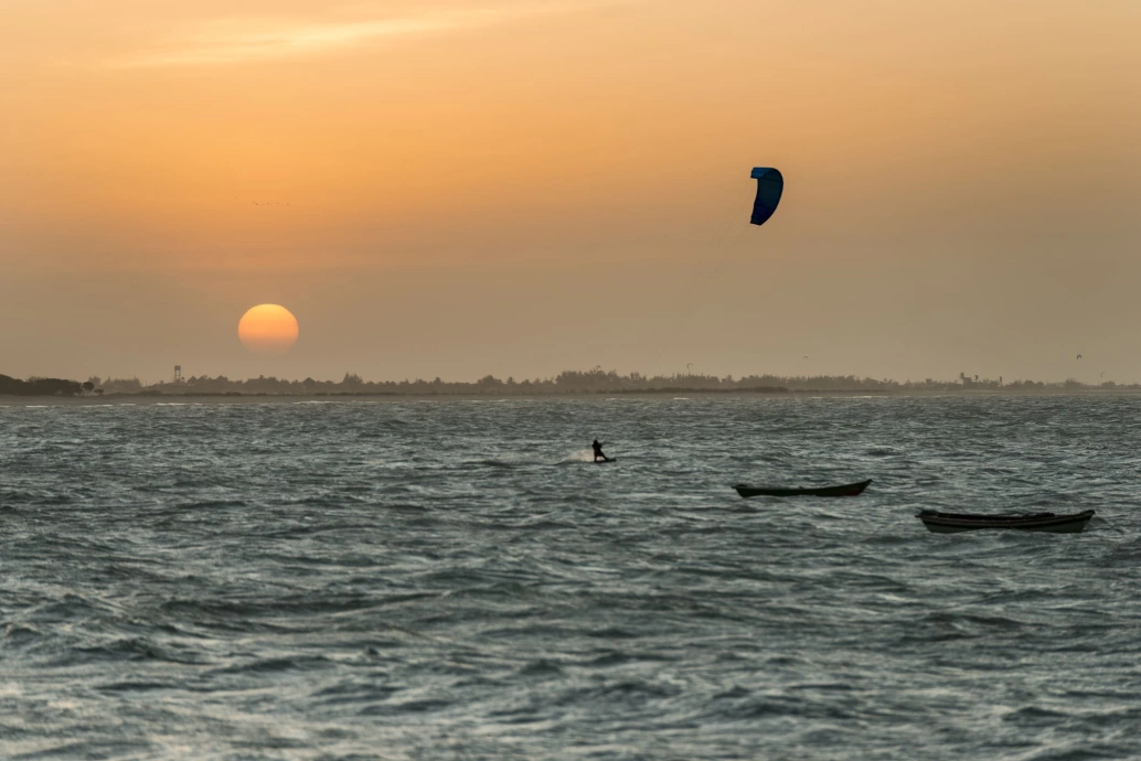 Mar no plano frontal e pôr do sol ao fundo. Um banhista pratica kitesurf próximo a dois pequenos barcos que repousam sobre a água