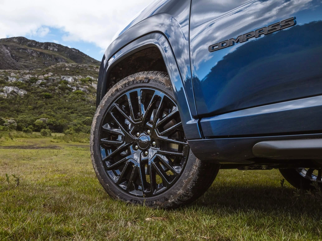 Roda dianteira de um Jeep Compass azul, que está estacionado em um gramado com montanhas ao fundo.