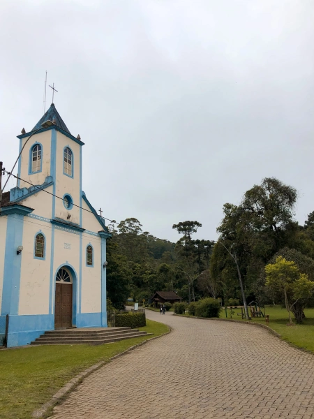 Vista de igreja clássica e simples, do período colonial no Brasil, junto à estrada calçada e natureza ao fundo, no município de Visconde de Mauá.