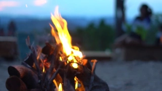 Destaque para uma fogueira acesa, feita com galhos de árvores. Mulher e paisagem desfocados ao fundo