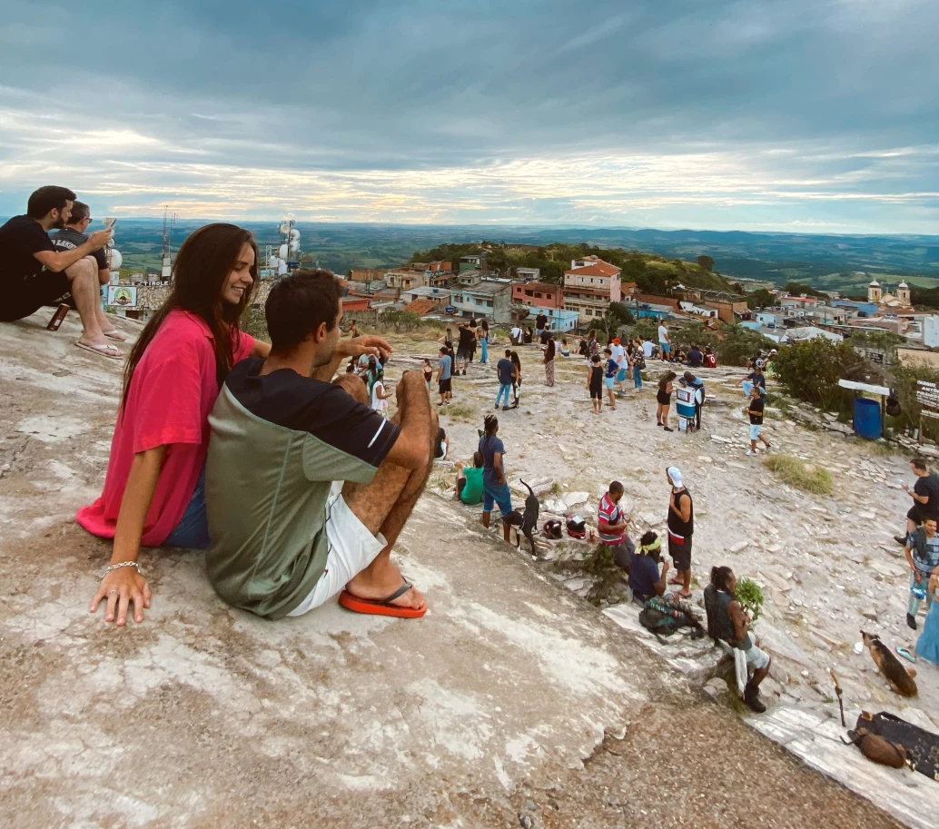 Homem e mulher sentados sobre uma pedra em um ponto alto da cidade de São Thomé das Letras. O local está cheio de pessoas e dá vista para algumas casas, além da paisagem montanhosa e céu parcialmente nublado ao fundo
