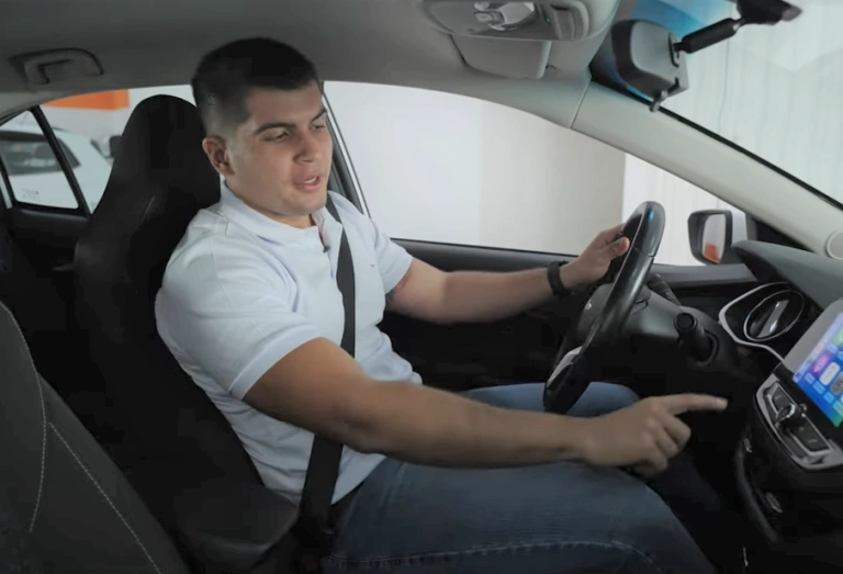 O influenciador Xenão está sentado no banco do motorista de um carro, com a mão esquerda ao volante e a outra apontando para a central multimídia.