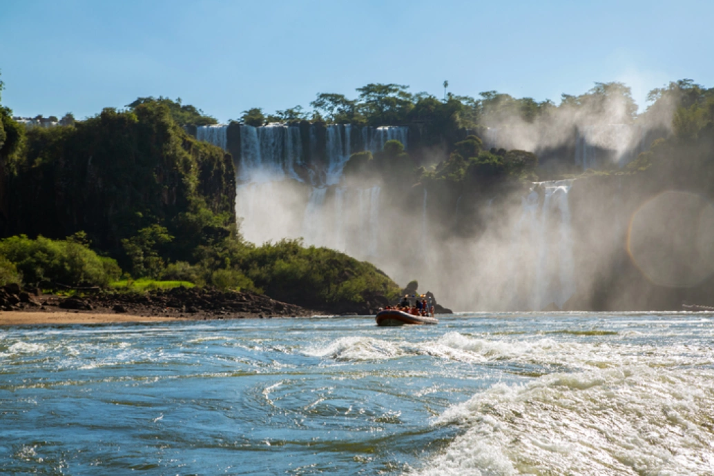 Fotografia ampla do Rio Iguaçu e suas quedas d’água ao fundo. Um bote com passageiros está ao centro da imagem. Vegetação ao lado esquerdo e entre as cataratas.