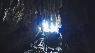 Homem em pé em meio às estruturas de uma caverna escura, com luz entrando por uma fenda e iluminando as estalactites no teto