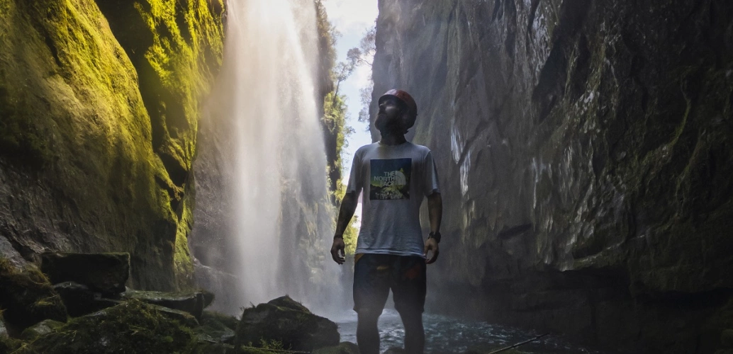 Fayson Merege em pé olhando para o alto na Cachoeira Véu da Noiva. Quedas d'água correm pelas paredes das rochas.