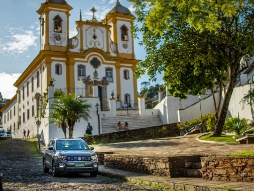 Num dia ensolarado, um carro estacionado em frente à uma igreja com arquitetura barroca nas cores branca e amarelo ouro com pessoas sentadas na escadaria em frente à praça.