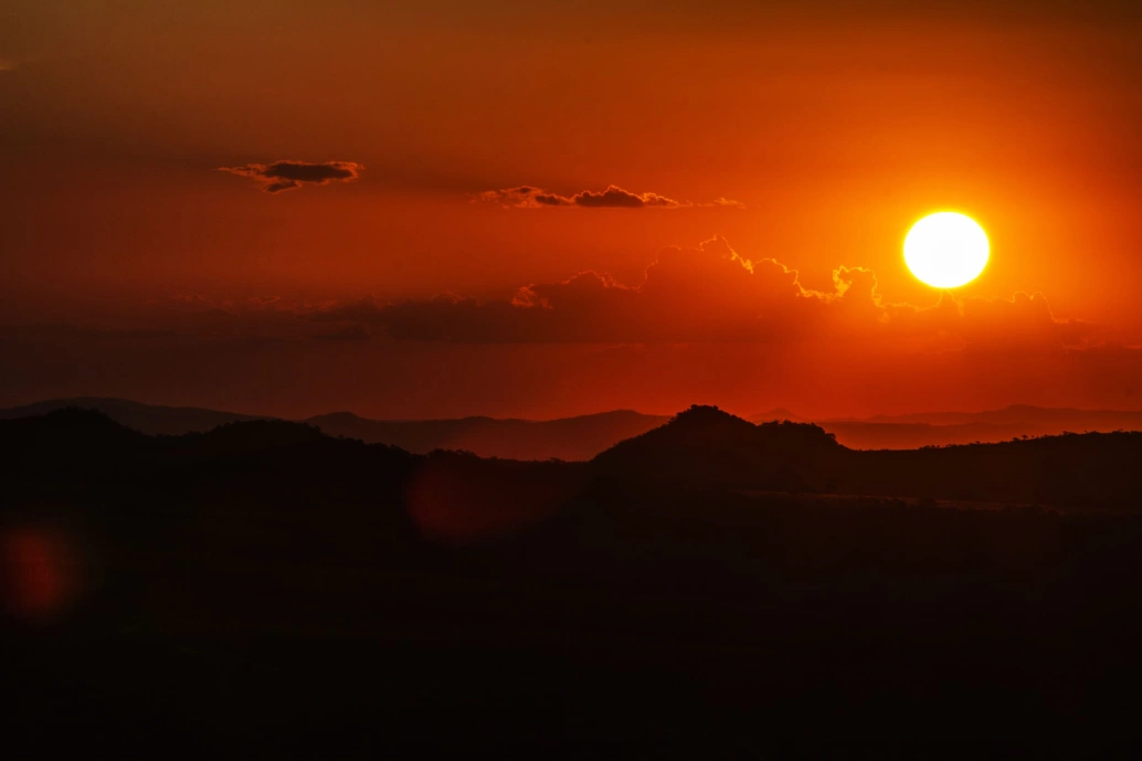 Pôr do sol visto do topo de uma montanha em Pirenópolis. O céu de coloração laranja intenso contrasta com a silhueta de outras montanhas. O sol aparece no canto direito da imagem.