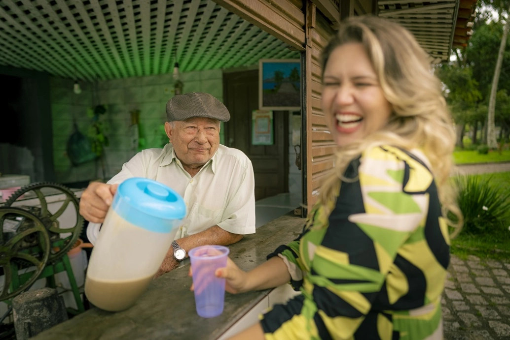 Mulher loira sorri enquanto recebe, no copo vazio de plástico que segura, caldo de cana de um homem de meia idade ao lado de moedor de cana manual. Uma bancada comercial os separam.