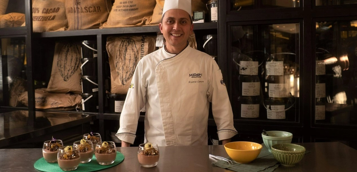 Chef Ricardo Campos sorrindo em frente a mesa contendo sobremesas