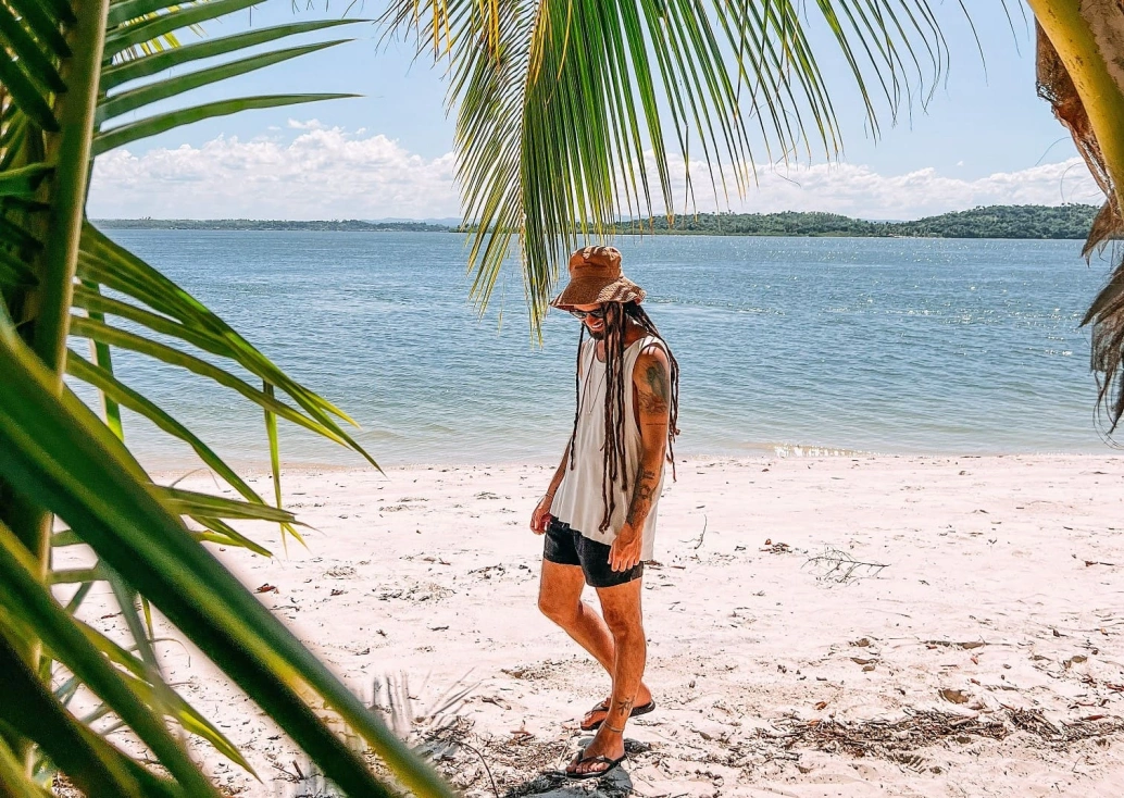 Homem em pé sob areia branca de praia deserta cercada por vegetação nativa