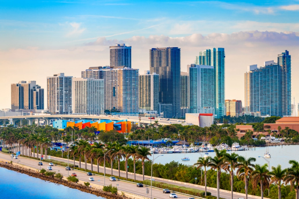Vista da cidade de Miami, Estados Unidos. Na porção inferior, uma rodovia com coqueiros em sua extensão e carros passando. No canto direito, o mar e várias embarcações ancoradas. Ao fundo, avista-se o céu e prédios enormes.