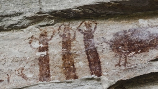 Parede rochosa com pinturas rupestres representando três pessoas em uma dança
