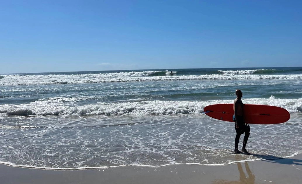 Praia em dia ensolarado, com ondas moderadas. À esquerda, um homem segura em baixo dos braços uma prancha de surf.